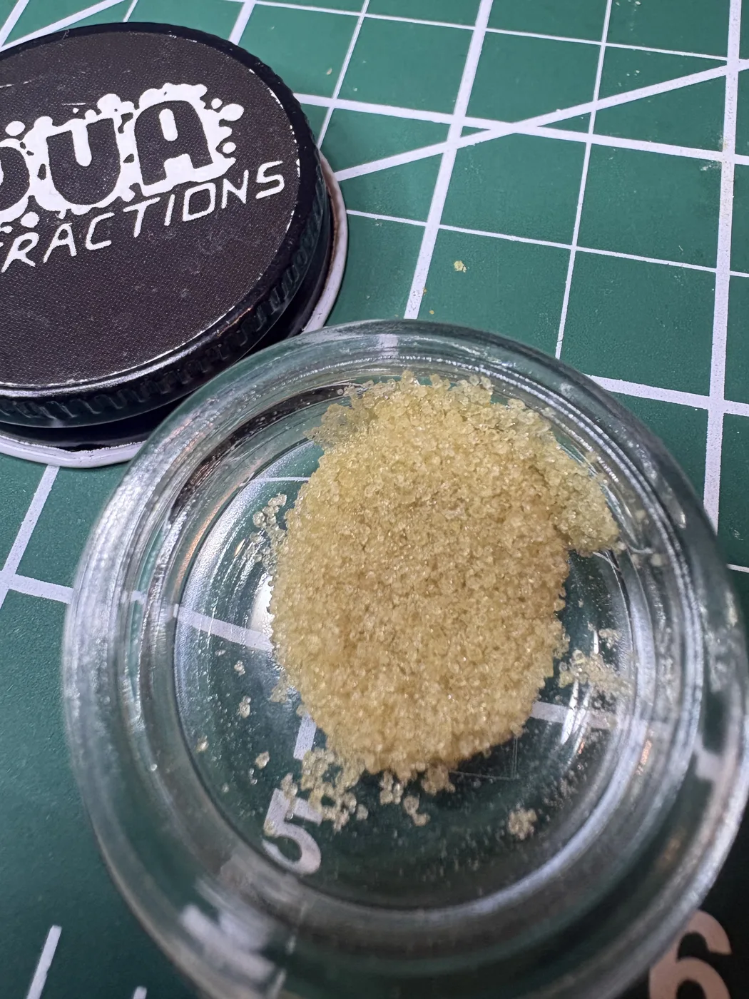 An open jar of loose, sandy, light-tan PUA Chem D hash beside its black PUA Extractions lid on a green cutting mat