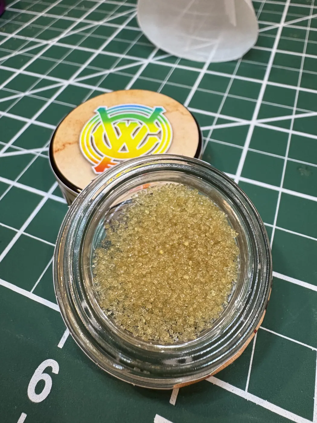 An open jar of sandy, light-gold West Coast Alchemy Vintage Z hash beside its rainbow West Coast Alchemy lid on a green cutting mat
