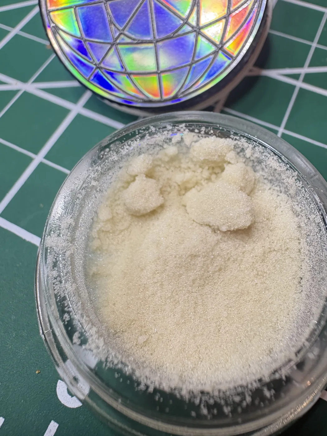 An open jar of bright white, powdery Wooksauce Grape Gas hash with a few soft chunks, the holographic Wooksauce lid behind it